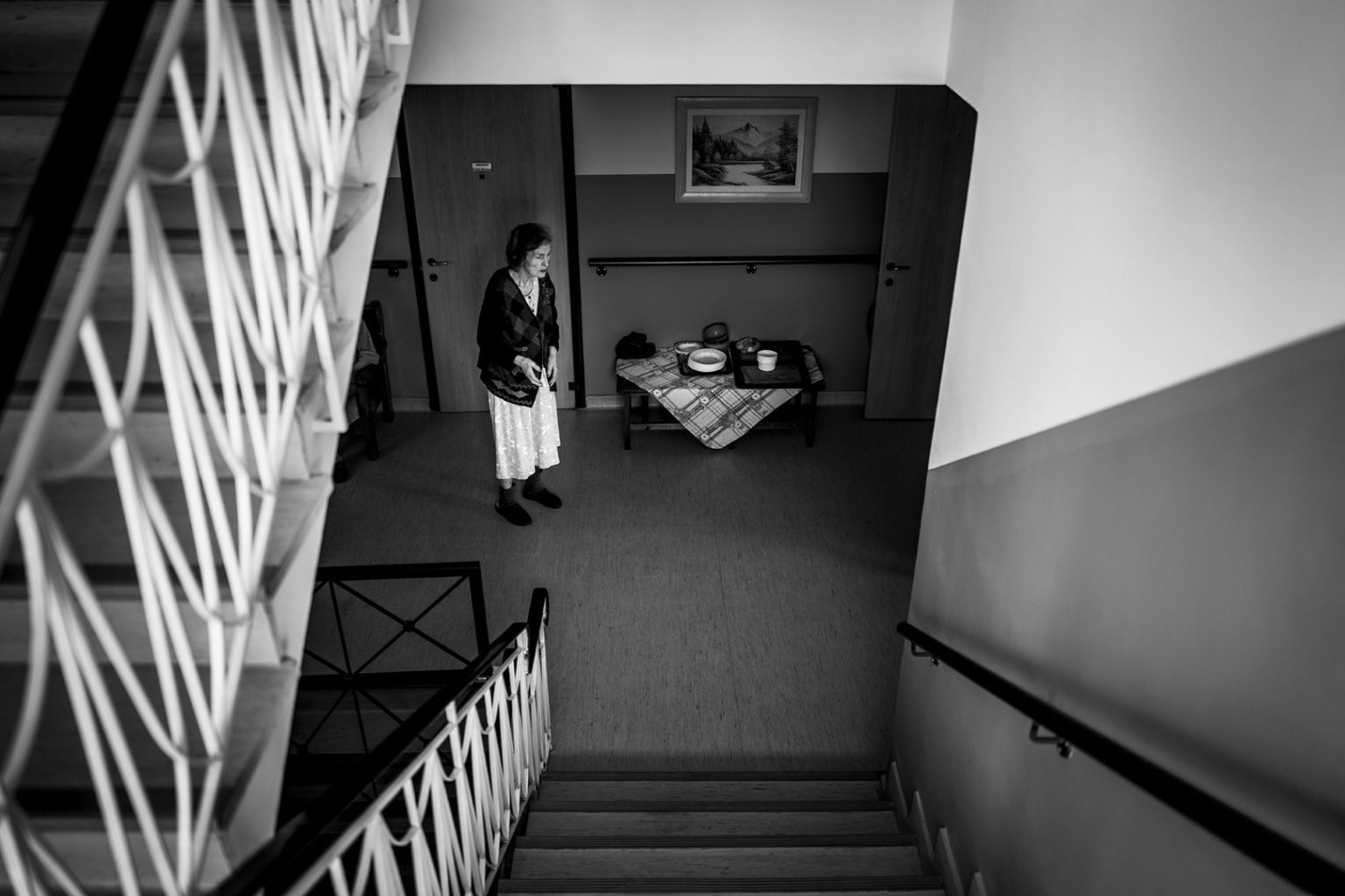 Μια ηλικιωμένη γυναίκα περιπλανιέται μόνη σε έναν διάδρομο του γηροκομείου, πλαισιωμένη από τη σιωπή. Η σκάλα στα αριστερά της μοιάζει να οδηγεί κάπου, αλλά εκείνη παραμένει καθηλωμένη στο παρόν. Στη μοναξιά της, η εικόνα της γίνεται σύμβολο ενός χρόνου που έχει σταματήσει, ενώ η άνοια σκιάζει την αντίληψή της, αφήνοντας πίσω μόνο αποσπασματικές αναμνήσεις.An elderly woman wanders alone in a nursing home corridor, surrounded by silence. The staircase on her left seems to lead somewhere, yet she remains anchored in the present. In her solitude, her image becomes a symbol of time standing still, as dementia clouds her perception, leaving behind only fragmented memories.