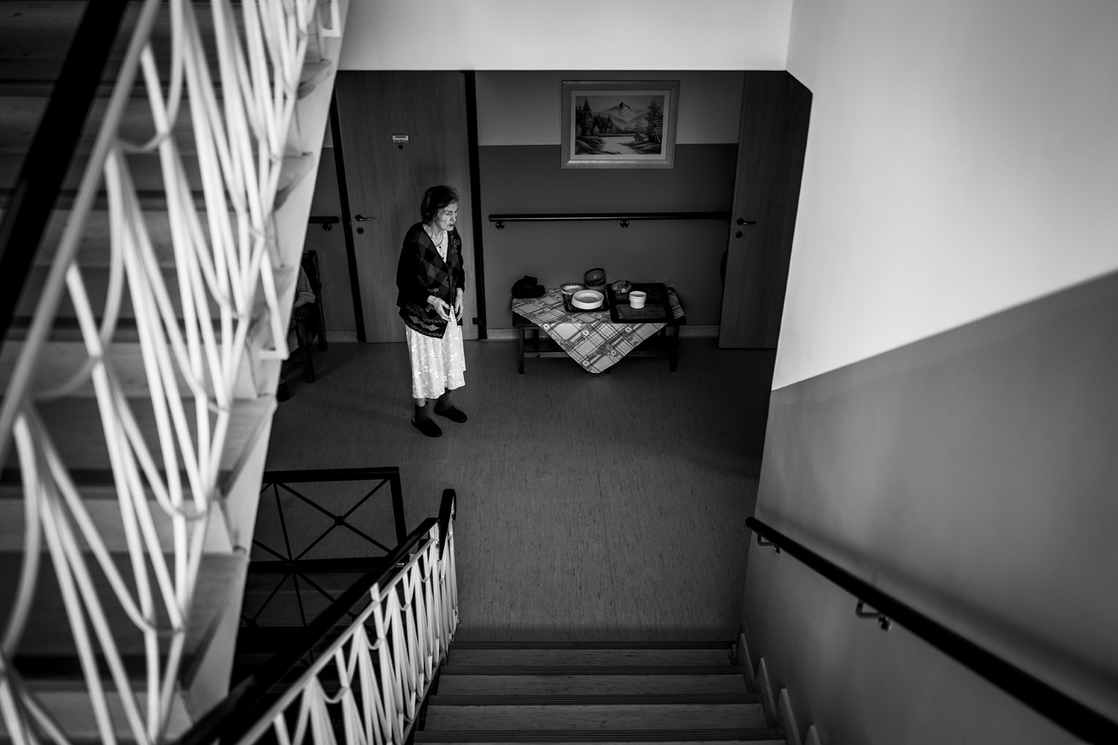 Μια ηλικιωμένη γυναίκα περιπλανιέται μόνη σε έναν διάδρομο του γηροκομείου, πλαισιωμένη από τη σιωπή. Η σκάλα στα αριστερά της μοιάζει να οδηγεί κάπου, αλλά εκείνη παραμένει καθηλωμένη στο παρόν. Στη μοναξιά της, η εικόνα της γίνεται σύμβολο ενός χρόνου που έχει σταματήσει, ενώ η άνοια σκιάζει την αντίληψή της, αφήνοντας πίσω μόνο αποσπασματικές αναμνήσεις.

An elderly woman wanders alone in a nursing home corridor, surrounded by silence. The staircase on her left seems to lead somewhere, yet she remains anchored in the present. In her solitude, her image becomes a symbol of time standing still, as dementia clouds her perception, leaving behind only fragmented memories.