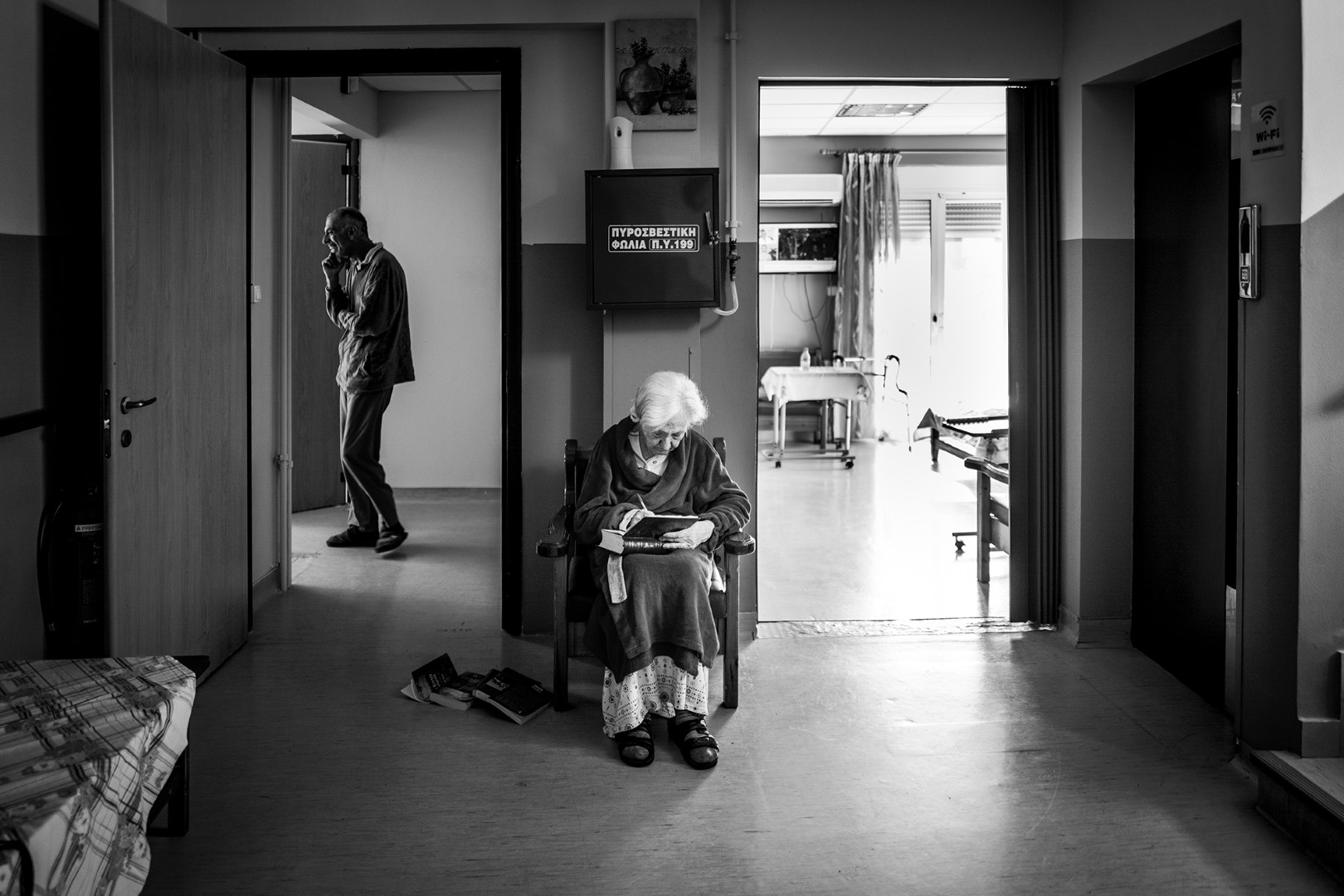 Μια ηλικιωμένη γυναίκα, πρώην ξεναγός, κάθεται μόνη σε μια ξύλινη καρέκλα, γράφοντας σε ένα βιβλίο. Τα πεσμένα βιβλία γύρω της μαρτυρούν την ανάγκη της να επικοινωνήσει με έναν κόσμο που μοιάζει να την έχει ξεχάσει. Πίσω της, ένας άλλος τρόφιμος στέκεται σιωπηλός, ενώ μια τηλεόραση παίζει μόνη της, τονίζοντας τη μοναξιά που γεμίζει το χώρο. Τα βιβλία της είναι το τελευταίο της παράθυρο προς την επικοινωνία και τη ζωή που κάποτε γνώριζε.

An elderly woman, a former tour guide, sits alone in a wooden chair, writing in a book. The scattered books around her reveal her yearning to connect with a world that seems to have forgotten her. Behind her, another resident stands in silence, while a television plays by itself, accentuating the solitude that fills the space. Her books are her last window to communication and the life she once knew.