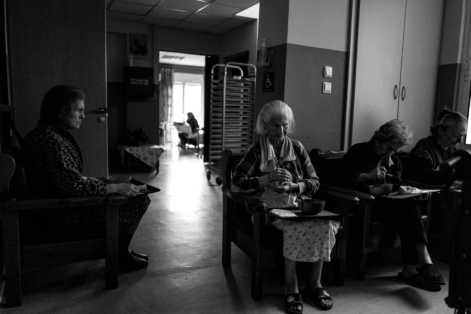 Τέσσερις ηλικιωμένες γυναίκες τρώνε το μεσημεριανό τους, καθισμένες η μία δίπλα στην άλλη, σιωπηλές. Στο βάθος, μια σκιά διακρίνεται αμυδρά, θυμίζοντας έναν χαμένο δεσμό. Κάποτε μοιράζονταν τραπέζια γεμάτα ζωντάνια, συζύγους, και παιδιά. Τώρα, μόνες, η συντροφικότητα περιορίζεται στην κοινή τους μοναξιά.

Four elderly women sit side by side, silently having their lunch. In the distance, a shadow faintly emerges, evoking memories of a lost connection. Once, they shared vibrant tables with spouses and children. Now, alone, their companionship is confined to their shared solitude.
